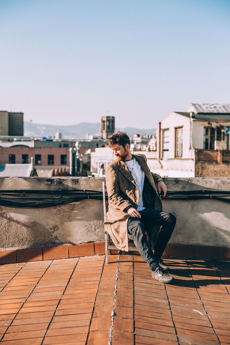 A stylishly dressed man is seated on a rooftop on a sunny day.