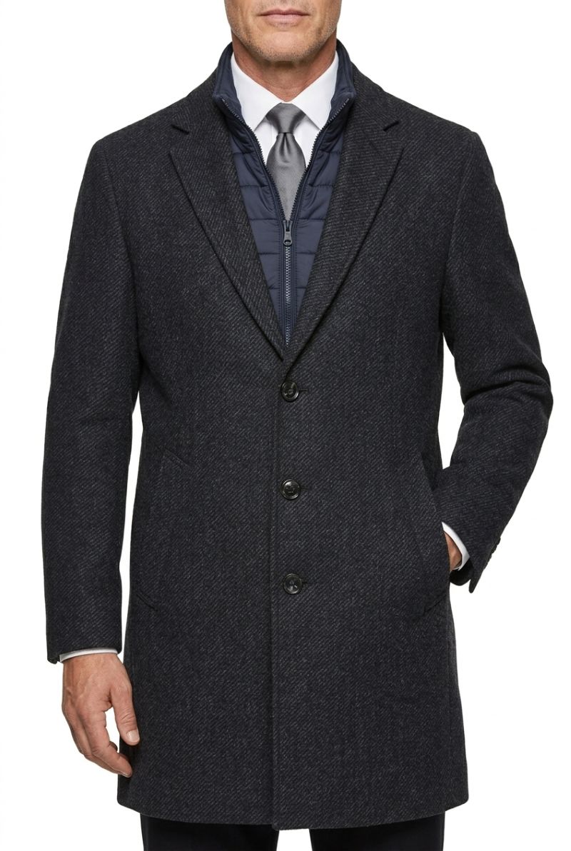 A man wearing a dark charcoal gray wool-blend overcoat over a navy quilted zip jacket, white dress shirt, and gray tie, shown from the waist up on a white background.