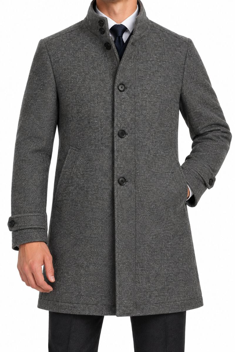 A man wearing a charcoal gray wool-blend coat with a high stand collar, three front buttons, and side pockets, paired with a white shirt and dark tie against a plain white background.