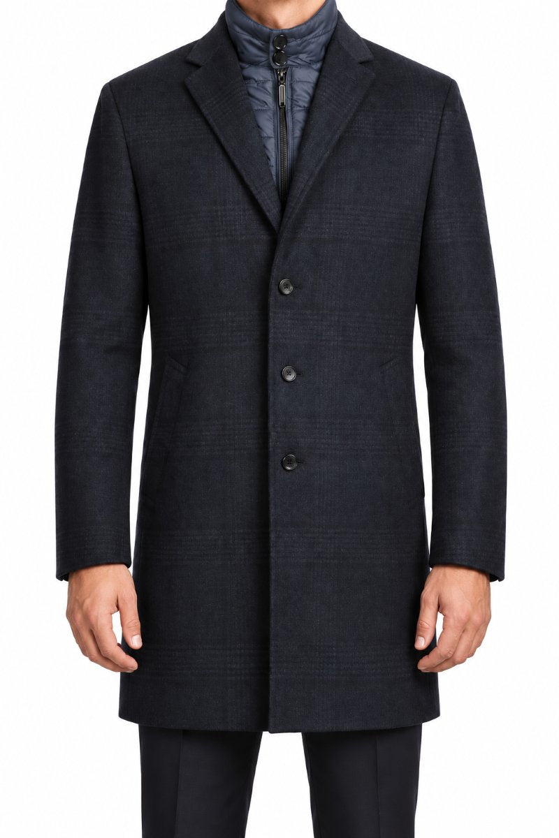 A man wearing a dark charcoal gray double-breasted overcoat with a subtle windowpane pattern, layered over a blue zippered jacket, shown against a plain white background.
