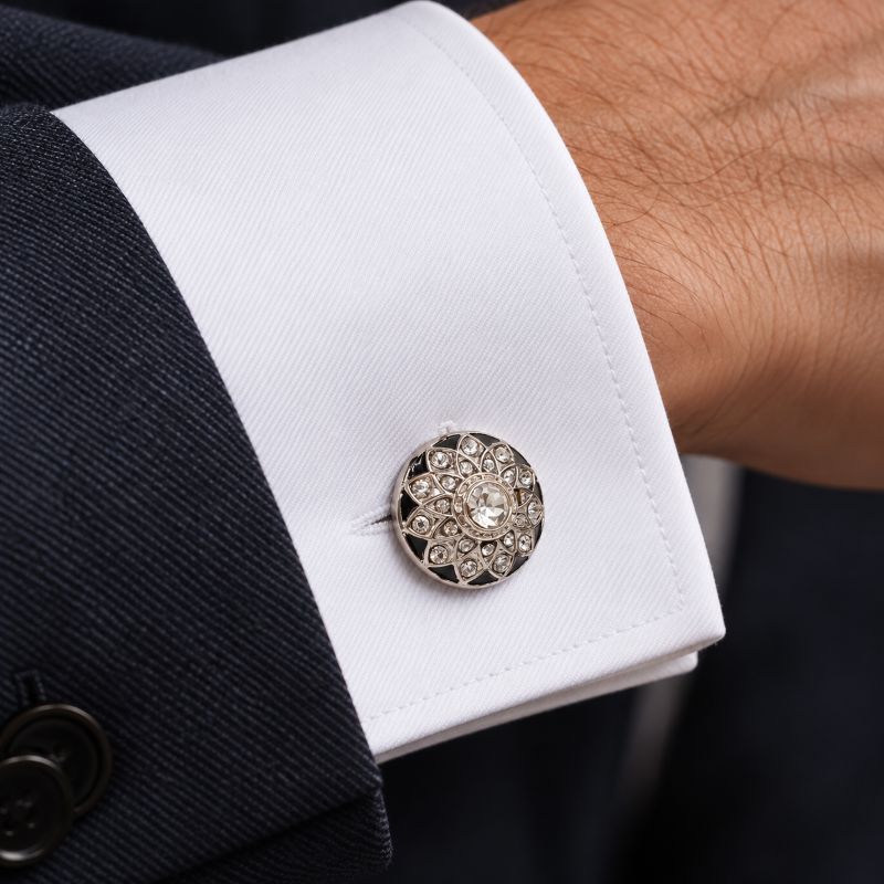 A close-up of a white dress shirt cuff with a decorative round silver cufflink, worn with a dark suit jacket on a person's wrist.