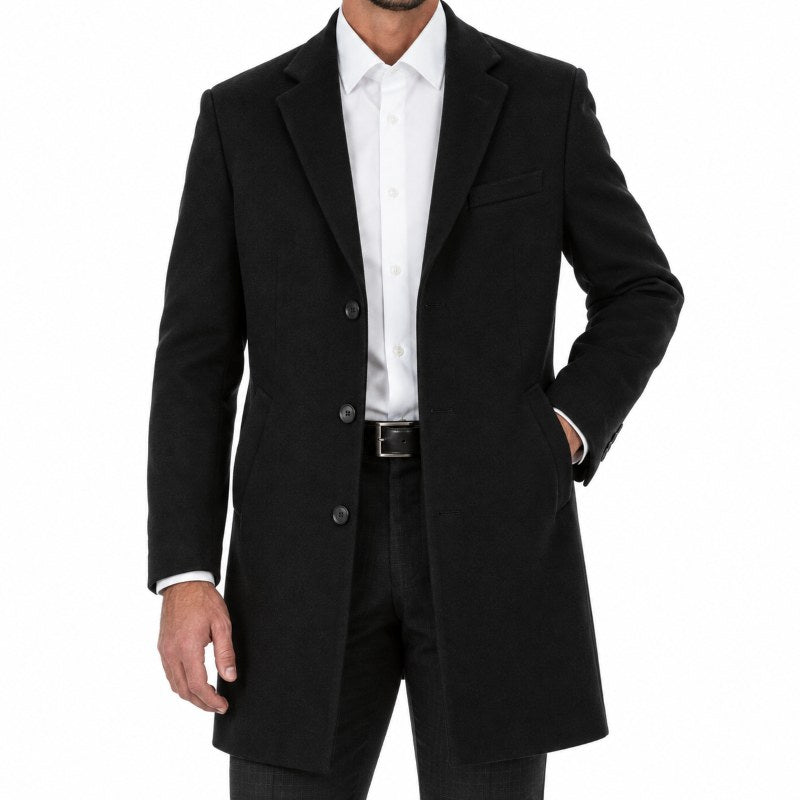 A man wearing a black mid-length overcoat over a white dress shirt and dark trousers, photographed against a plain white studio background.