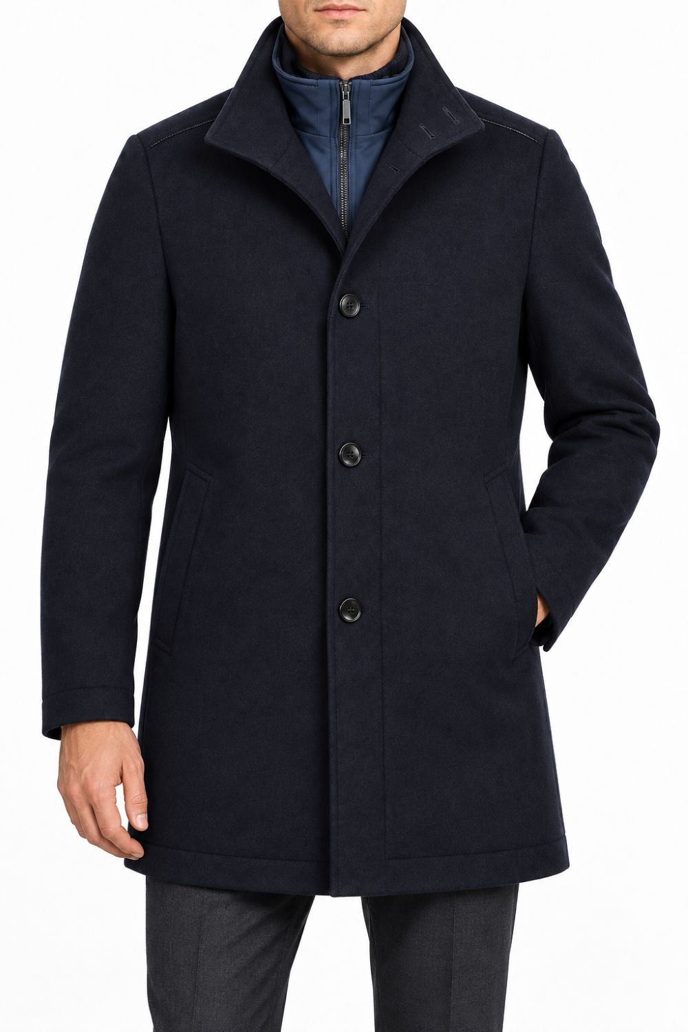 A man wearing a dark navy blue textured overcoat over a zippered navy jacket, white shirt, and dark tie, shown from the neck down on a plain white background.