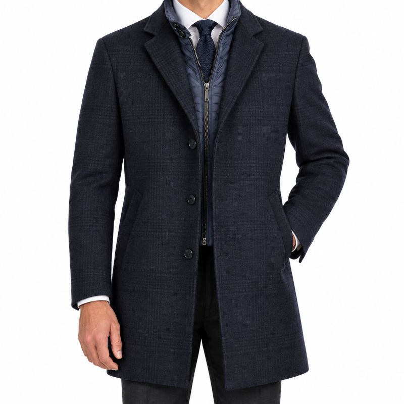 A man wearing a dark blue textured overcoat over a zippered navy jacket, white shirt, and dark tie, shown from the neck down on a plain white background.