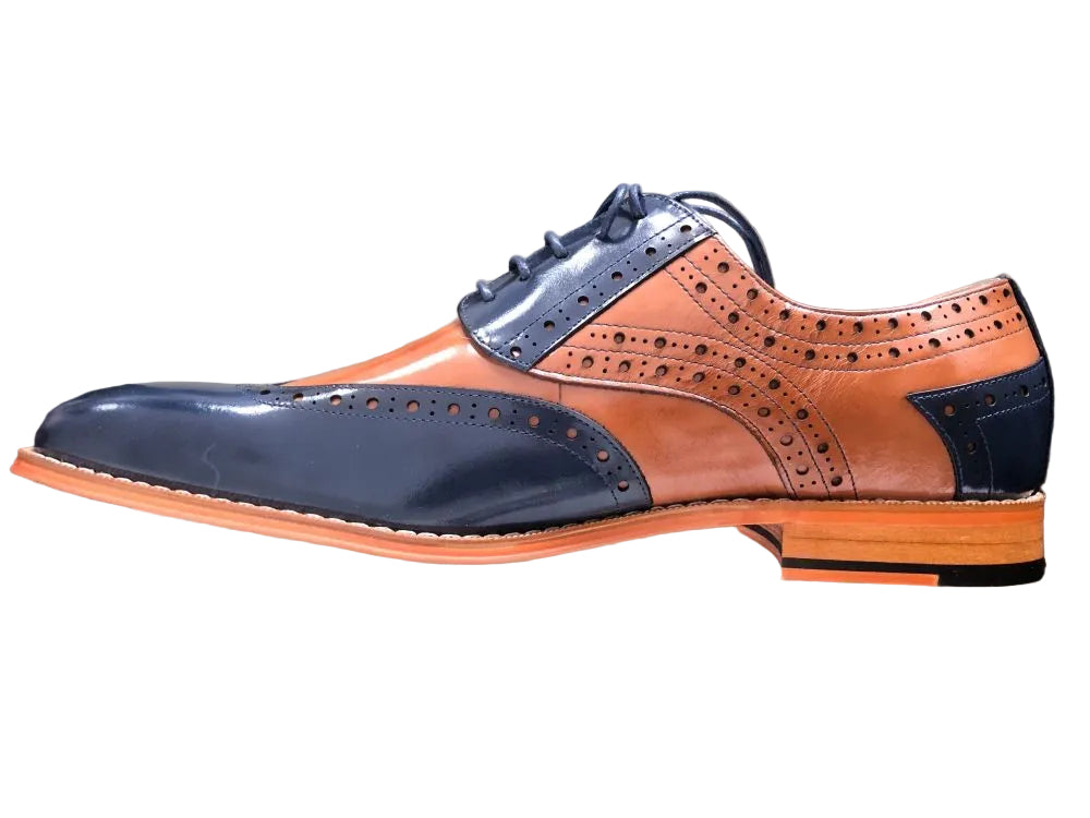 A men's wingtip oxford shoe with a two-tone color-blocked design, featuring dark navy blue leather on the toe, laces, and heel, and cognac brown leather on the quarter panels and vamp. The shoe is positioned horizontally on top of a light beige rectangular display block against a black background.