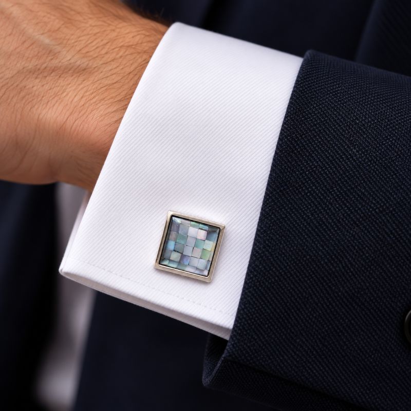 A square cufflink with a mosaic iridescent shell inlay and a gold-tone frame, shown fastened to a white dress shirt cuff on a person wearing a dark suit jacket.