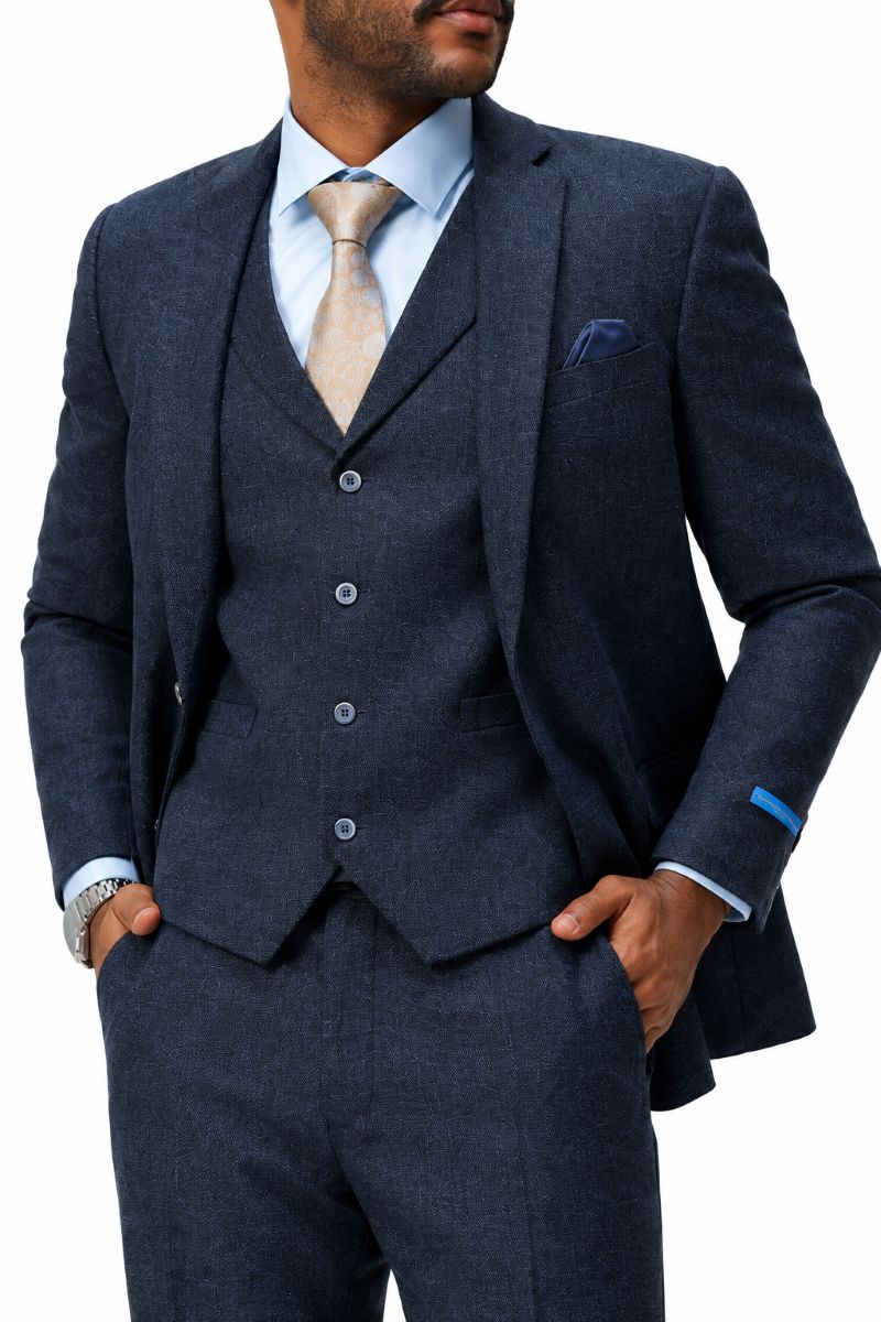 A man wearing a dark navy blue three-piece suit with a matching vest and trousers, styled with a light blue dress shirt, a beige tie, and a blue pocket square, against a plain white background.