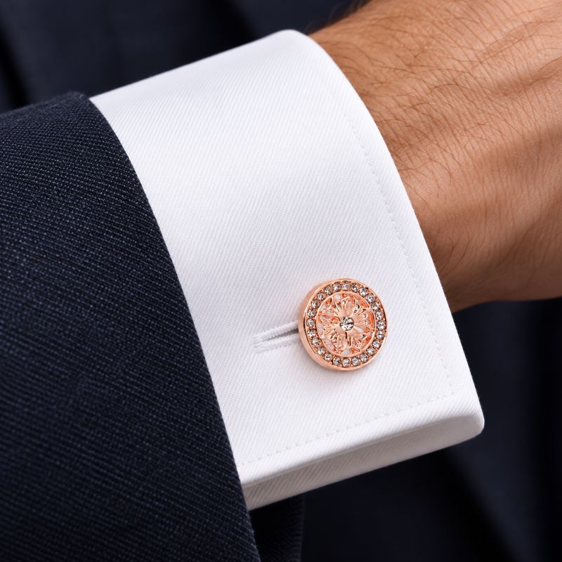 A close-up of a white dress shirt cuff with a dark navy suit jacket sleeve, featuring a single rose gold cufflink adorned with small clear crystals.