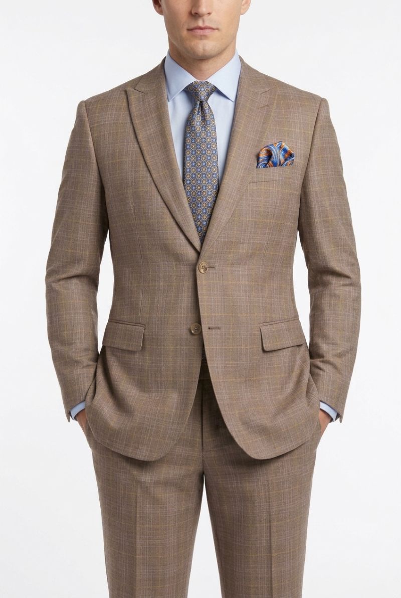 A man wearing a brown and beige plaid suit with a light blue dress shirt, patterned tie, and matching pocket square, photographed against a plain white studio background.
