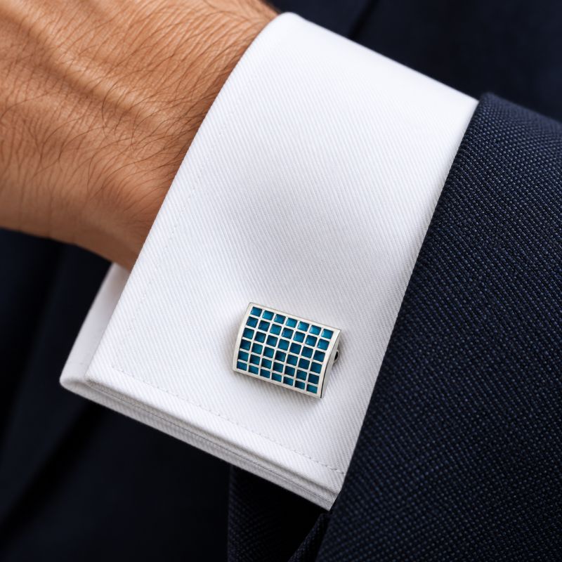 A close-up of a white dress shirt cuff with a rectangular cufflink featuring a blue grid pattern on a silver-toned base, worn with a dark navy suit jacket.