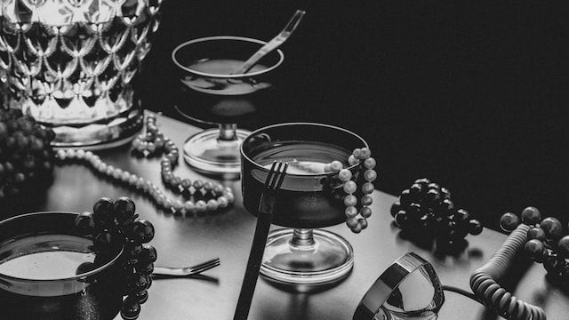 Black and white photo of elegant cocktail glasses with pearls draped over them, grapes on the table, and a crystal lamp creating a vintage, glamorous Gatsby party atmosphere.
