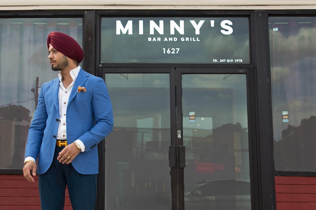 a man with a turban, wearing a stylish blue blazer over a white shirt, dark blue trousers, and a prominent gold-colored belt buckle