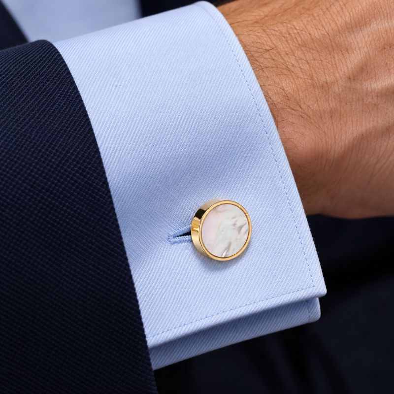 A close-up of a light blue dress shirt cuff with a single round gold cufflink featuring an off-white mother-of-pearl face, worn with a dark suit jacket on a person's wrist.