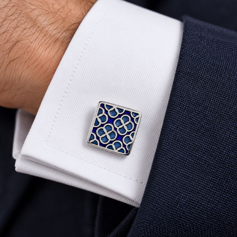 A square silver-toned cufflink with a blue and white geometric interlocking pattern, shown fastened on a white dress shirt cuff worn with a dark navy suit jacket.