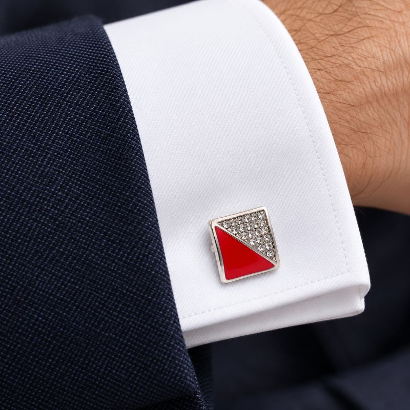 A close-up of a white dress shirt cuff worn on a wrist, featuring a square silver-toned cufflink with a red and silver geometric design.